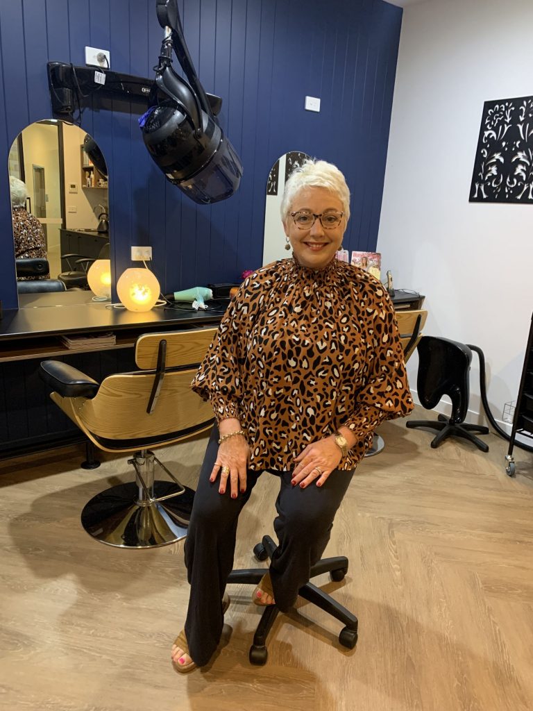 Hairstylist Lorrae sits on a chair wearing a leopard print shirt in the Brookland hair and beauty salon in front of a blue wall.