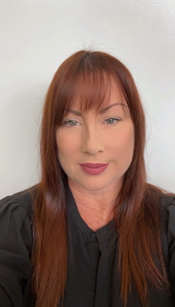 Hairstylist Kaylene holds phone and takes selfie in a black blouse with light auburn hair.