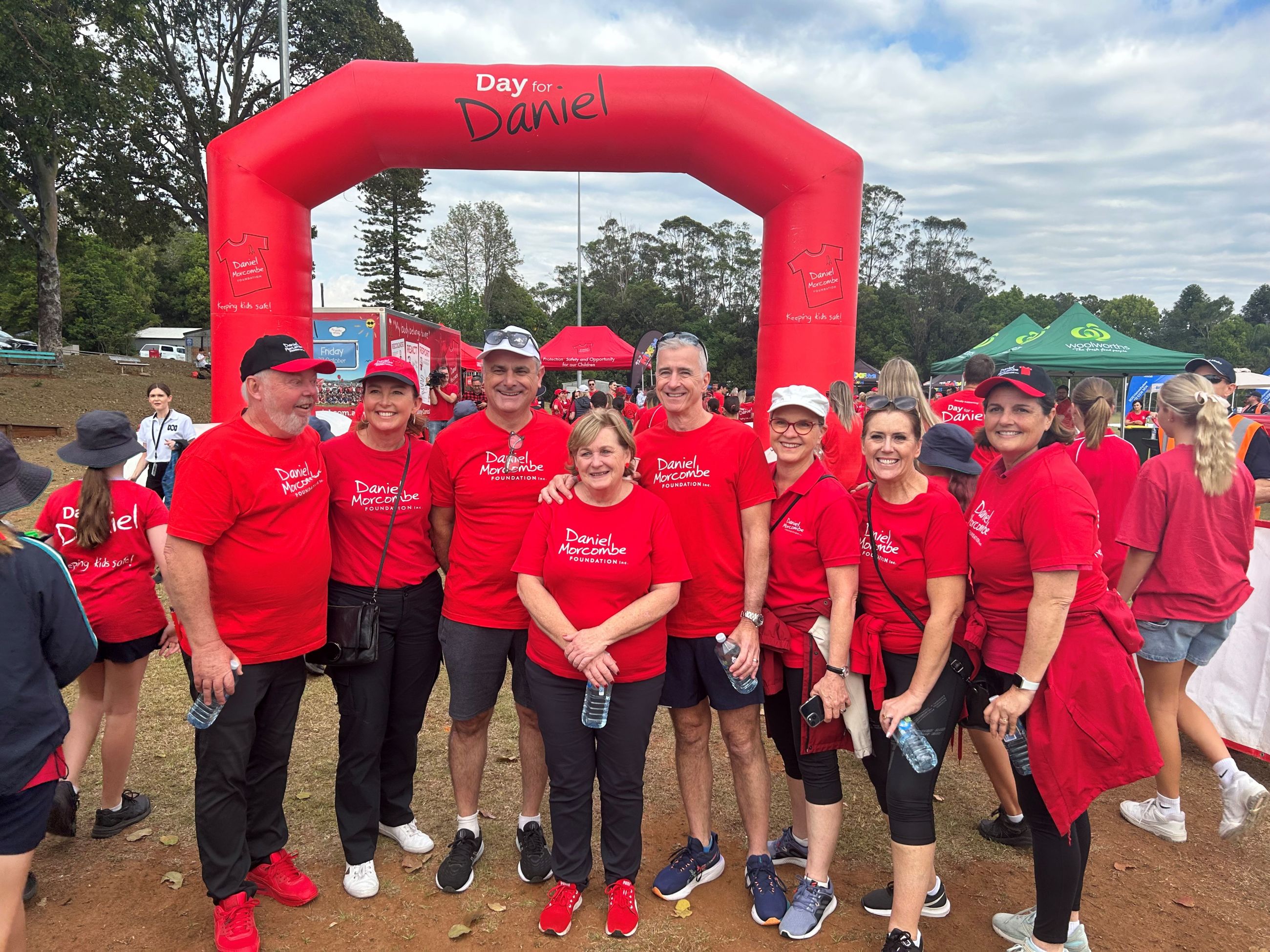 Group of Reside staff with Morcombe family for the Day for Daniel walk, group wear red t-shirts and stand under a large arch.
